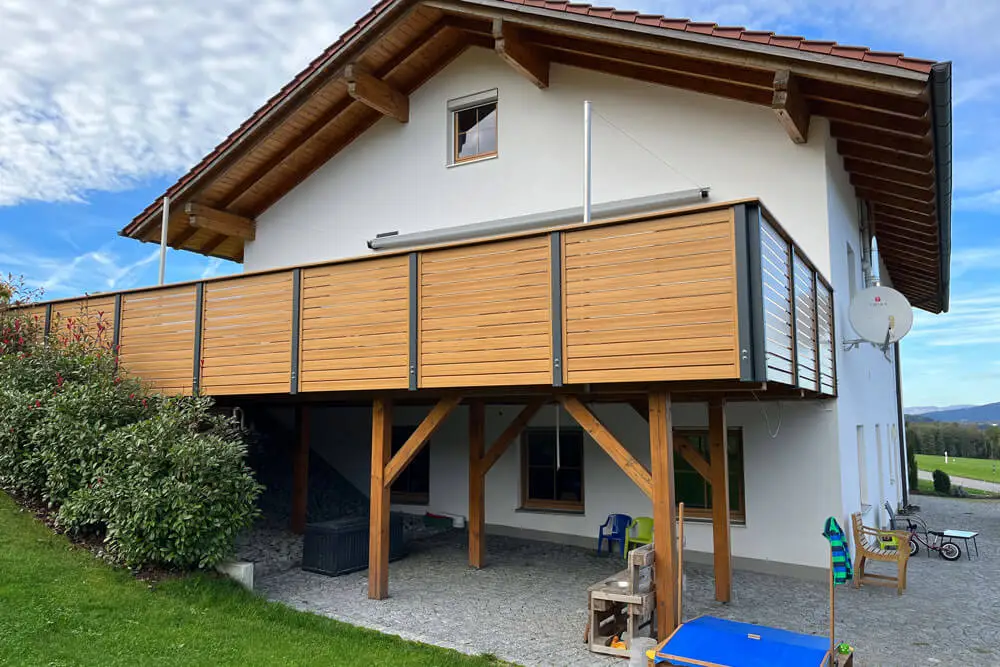 Kunststoff-Balkon Modell Wagrain mit Lärche-Dekor und anthrazitfarbenen Alu-Balustern an weißem Haus mit Holzdach und Holzfstützen unter Balkon