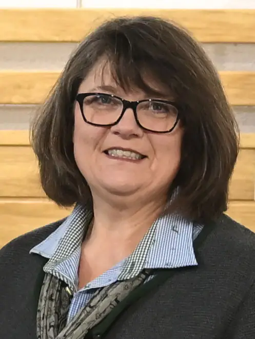Teammitglied mit schulterlangem braunem Haar, trägt ein blau-weiß gestreiftes Hemd, einen grauen Pullover und ein gemustertes Halstuch.