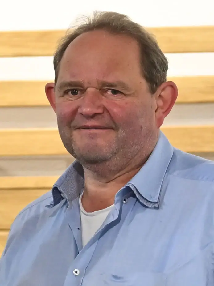 Mann mit grauem Haar trägt hellblaues Hemd über weißem T-Shirt, vor Holzlamellenwand stehend.