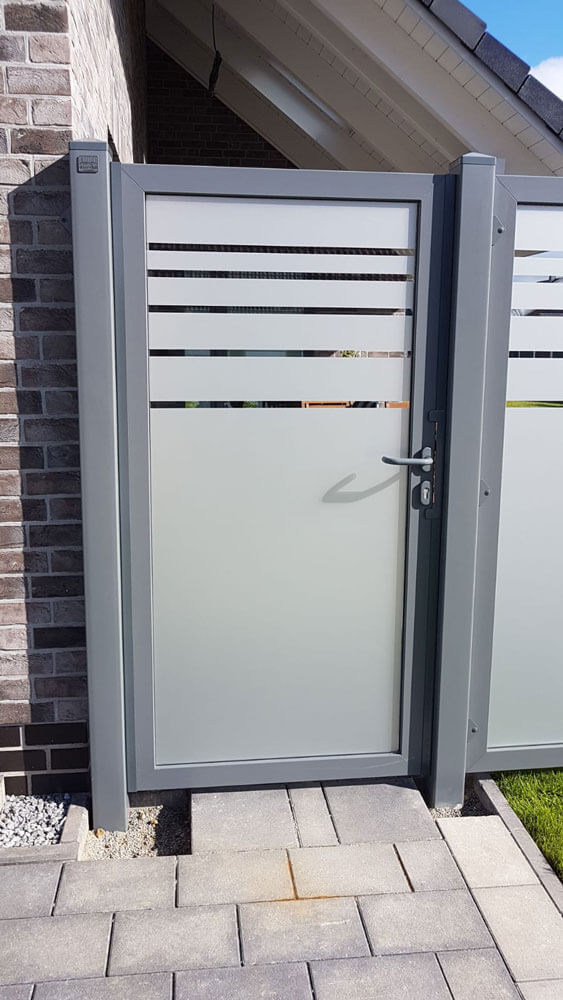 Sichtschutz-Einzeltor Serie 61, Glasfüllung mit folierten Streifen, eingebaut zwischen grauen Pfosten neben einem Haus mit Ziegelfassade