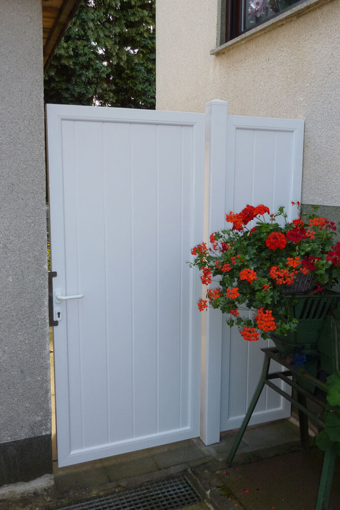 Weißes Sichtschutz-Einzeltor Serie 21 zwischen Hauswand und Mauer, neben Topf mit roten Blumen