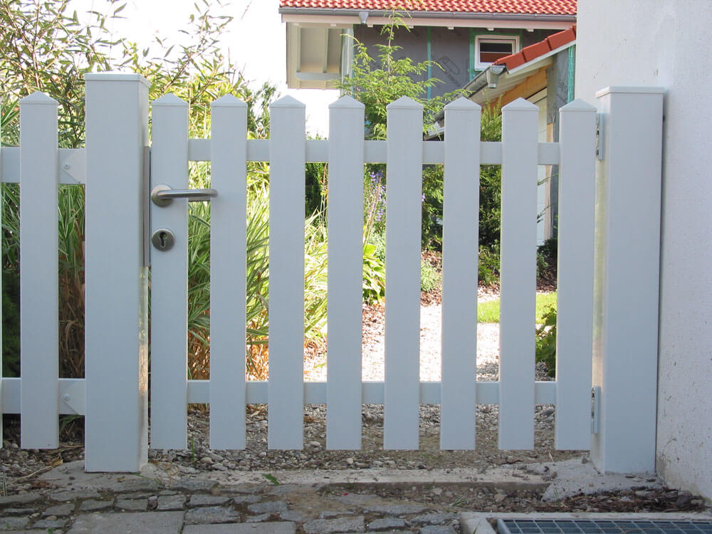 Einzeltor Saaletal aus weißem Kunststoff mit senkrechten Latten und Metallgriff, an weißer Hauswand befestigt, vor einem grauen Haus mit rotem Ziegeldach