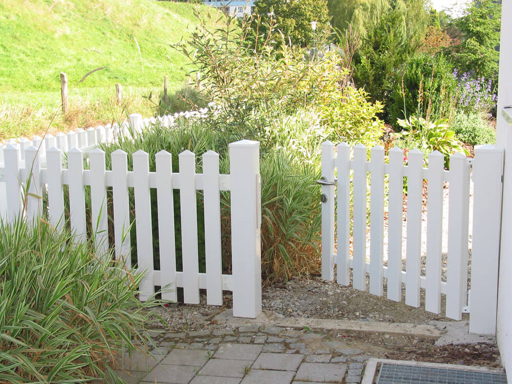 Einzeltor Saaletal aus weißem Kunststoff mit senkrechten Latten und Metallgriff, eingebaut in Gartenzaun mit Pflanzen im Hintergrund