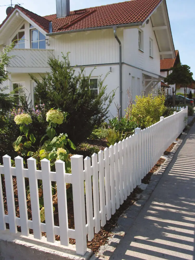Weißer Kunststoff-Zaun Modell Saaletal mit Spitzkappen umgibt einen Garten vor einem weißen Wohnhaus