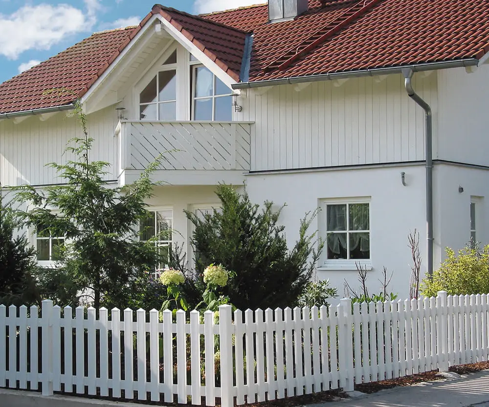 Weißer Kunststoff-Zaun Modell Saaletal mit Spitzkappen umgibt einen Garten vor einem Haus mit rotem Dach und Balkon