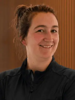 Freundlich lächelnde Frau mit zusammengebundenem Haar und verschränkten Armen trägt schwarze Jacke mit rotem Emblem am linken Ärmel und blaue Jeans vor Holzlamellenwand